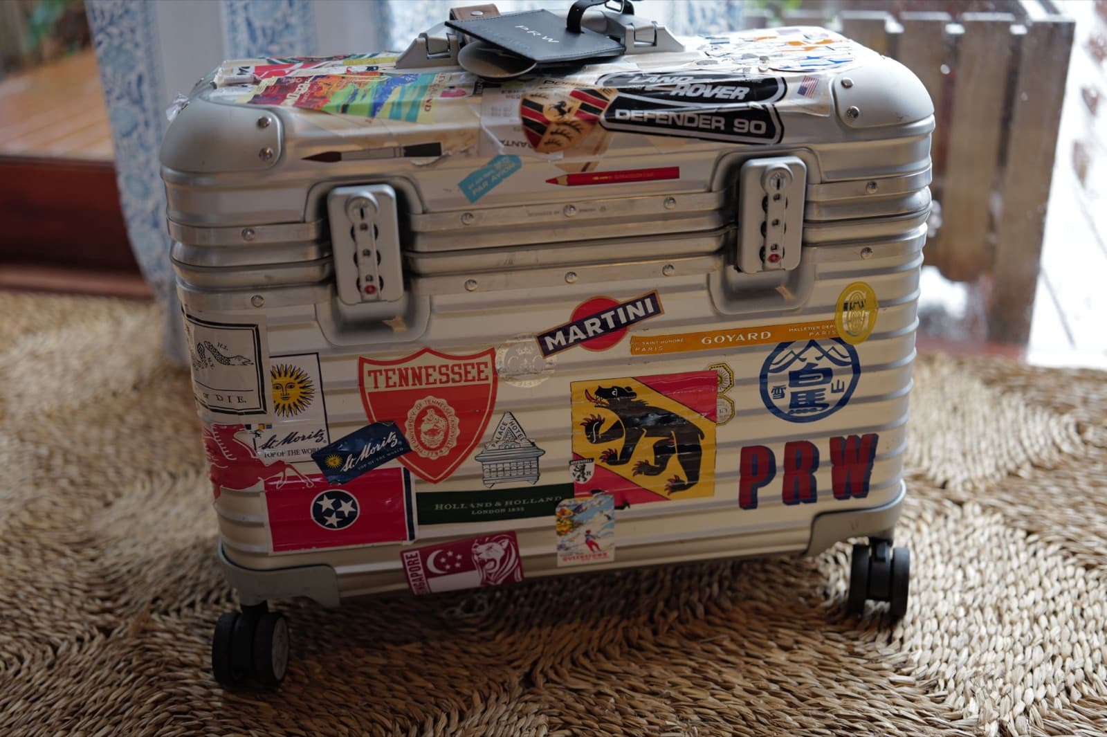 A silver aluminium Rimowa Pilot case densely covered in travel stickers, with a monogrammed leather luggage tag on top.