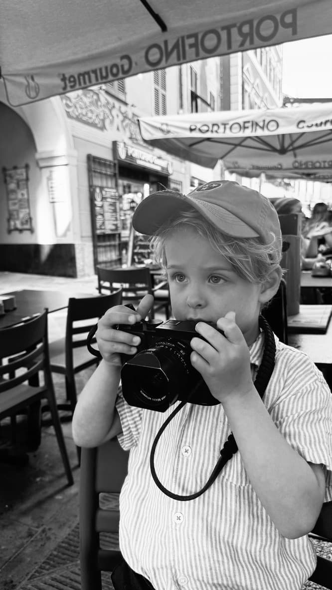 A child in a cap holds a Leica Q2 to his chin at an outdoor café table under a Portofino Gourmet awning.